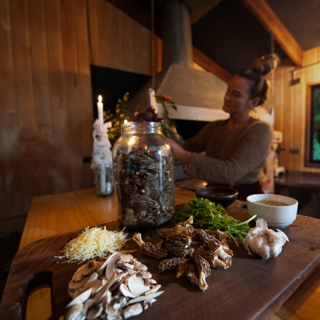 Escena de cocina con varios ingredientes sobre una tabla de cortar de madera, incluyendo una jarra de vidrio llena de hongos secos, ajo, queso rallado, champiñones en rodajas y hierbas frescas. Al fondo, una persona está de pie cerca de una chimenea o estufa, con velas encendidas y flores decorativas, creando un ambiente acogedor y rústico.