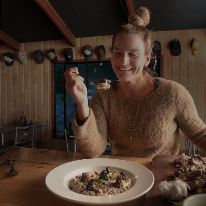 Persona sentada, sosteniendo un tenedor con un bocado de risotto de setas cerca de la boca. Lleva un suéter de lana y un collar. En la mesa hay un plato de risotto con setas y hierbas frescas, junto a una tabla de cortar con un diente de ajo y setas. Al fondo, se ve una pared de madera con varias gorras colgadas y una ventana que da al exterior oscuro.