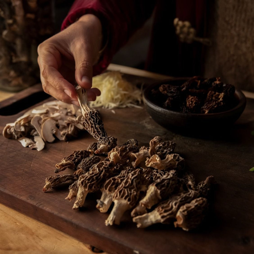 Persona recolectando hongos morilla de una tabla de cortar en una cocina, con hongos rebanados y queso rallado en el fondo.