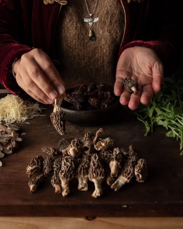 Persona preparando hongos morilla frescos en una tabla de cortar, rodeada de queso rallado, hojas verdes y más hongos en un tazón.