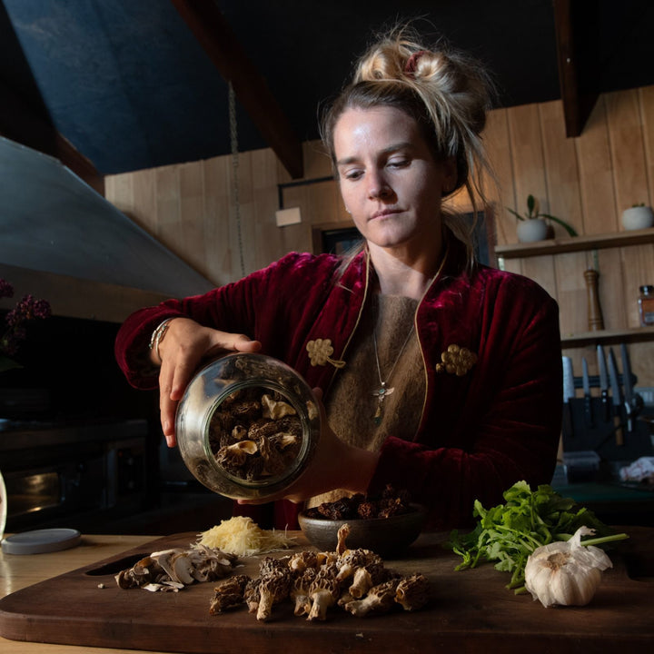 Persona vertiendo hongos secos de un frasco de vidrio sobre una tabla de cortar en una cocina de diseño rústico. En la tabla de cortar hay varios ingredientes, incluyendo hongos frescos, ajo, y hojas verdes. La persona lleva una chaqueta de terciopelo rojo y un collar. Los estantes de madera y los utensilios de cocina son visibles en el fondo.