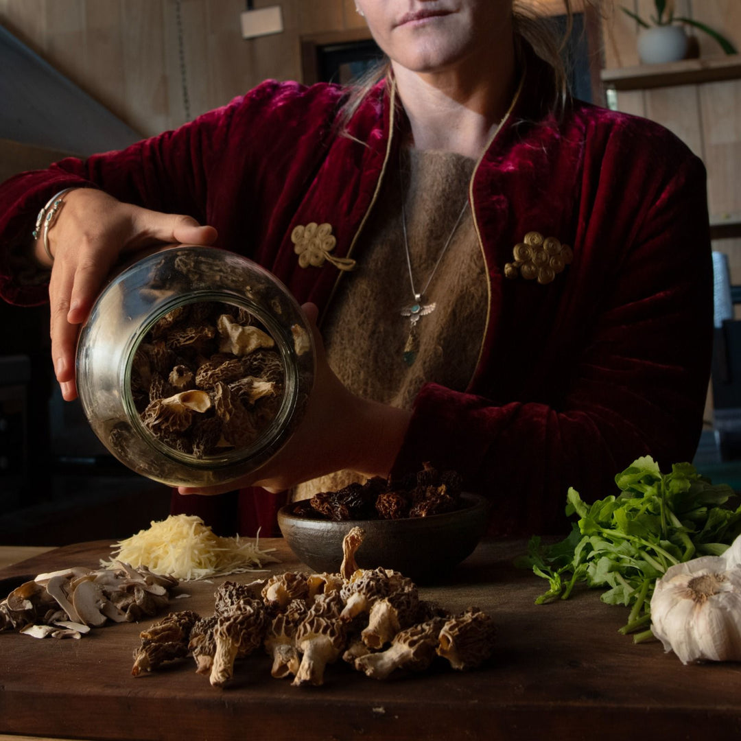 Persona vertiendo hongos secos de un frasco de vidrio sobre una mesa de madera en una cocina. En la mesa hay varios ingredientes, incluyendo más hongos, ajo, queso rallado y hojas verdes. La persona lleva una chaqueta de terciopelo rojo con adornos dorados y un collar.
