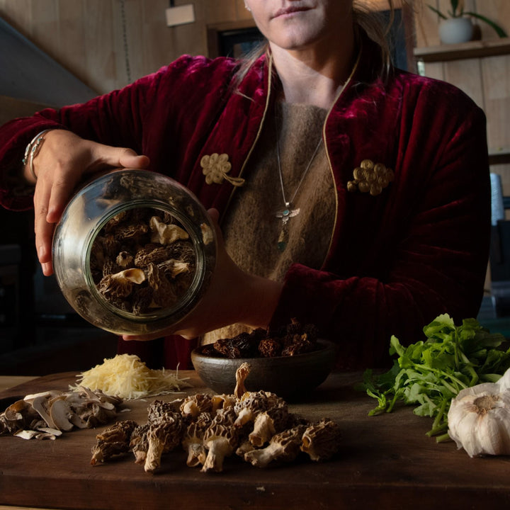 Persona vertiendo hongos secos de un frasco de vidrio sobre una mesa de madera en una cocina. En la mesa hay varios ingredientes, incluyendo más hongos, ajo, queso rallado y hojas verdes. La persona lleva una chaqueta de terciopelo rojo con adornos dorados y un collar.