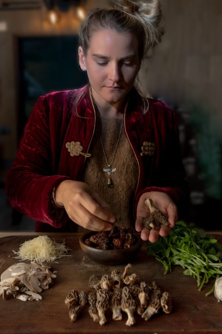 Una persona con una chaqueta roja y un suéter marrón está preparando ingredientes en una mesa de madera. En la mesa hay una variedad de hongos, incluyendo colmenillas frescas y secas, así como hongos en rodajas. También hay un montón de hojas verdes y un poco de queso rallado. La persona sostiene una colmenilla fresca en una mano y parece estar colocando hongos secos en un tazón de madera con la otra mano.