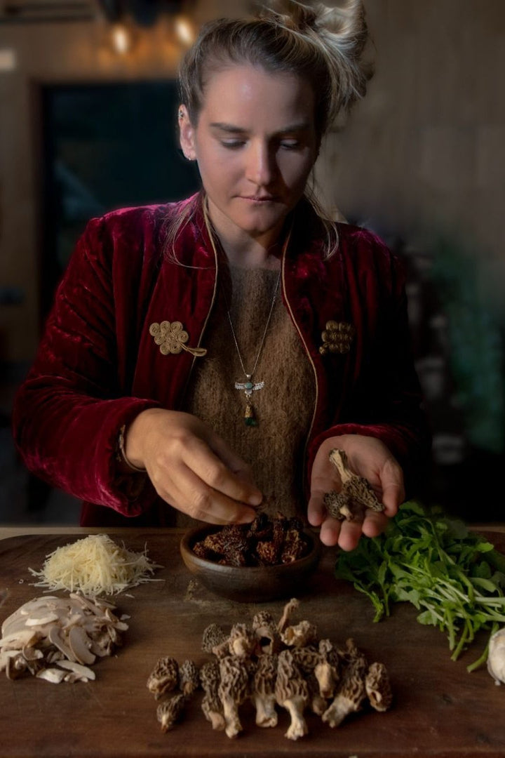 Una persona con una chaqueta roja y un suéter marrón está preparando ingredientes en una mesa de madera. En la mesa hay una variedad de hongos, incluyendo colmenillas frescas y secas, así como hongos en rodajas. También hay un montón de hojas verdes y un poco de queso rallado. La persona sostiene una colmenilla fresca en una mano y parece estar colocando hongos secos en un tazón de madera con la otra mano.