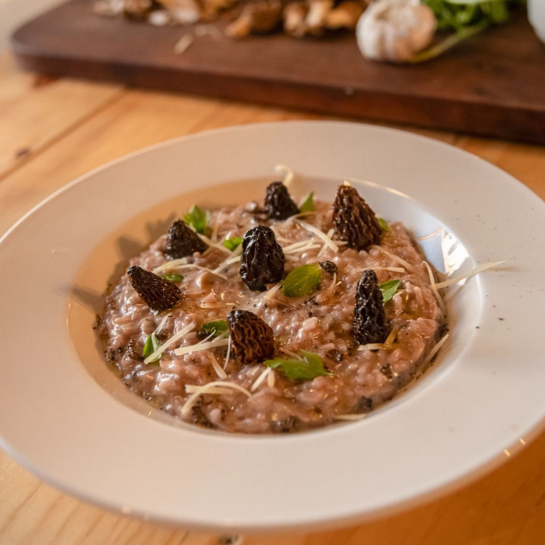 Un plato de risotto cremoso adornado con setas morillas, hojas verdes y queso rallado, servido en un plato blanco sobre una mesa de madera.