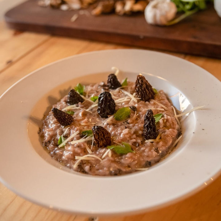 Un plato de risotto cremoso adornado con setas morillas, hojas verdes y queso rallado, servido en un plato blanco sobre una mesa de madera.