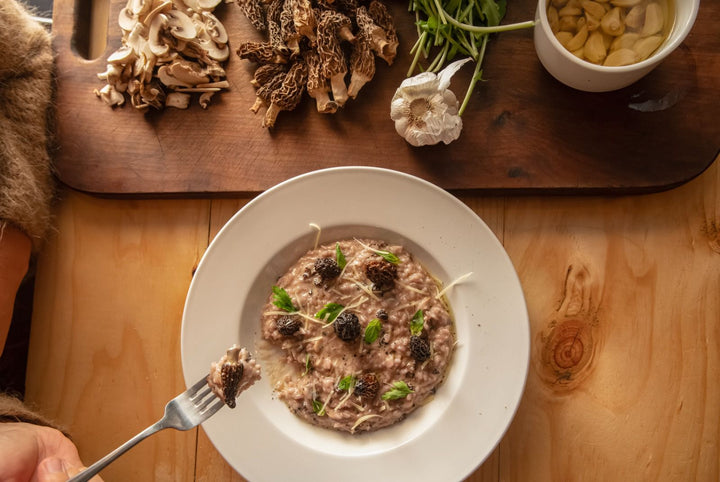Un plato de risotto cremoso con setas, decorado con hierbas frescas y ajo, junto a una tabla de cortar con más setas y hierbas.